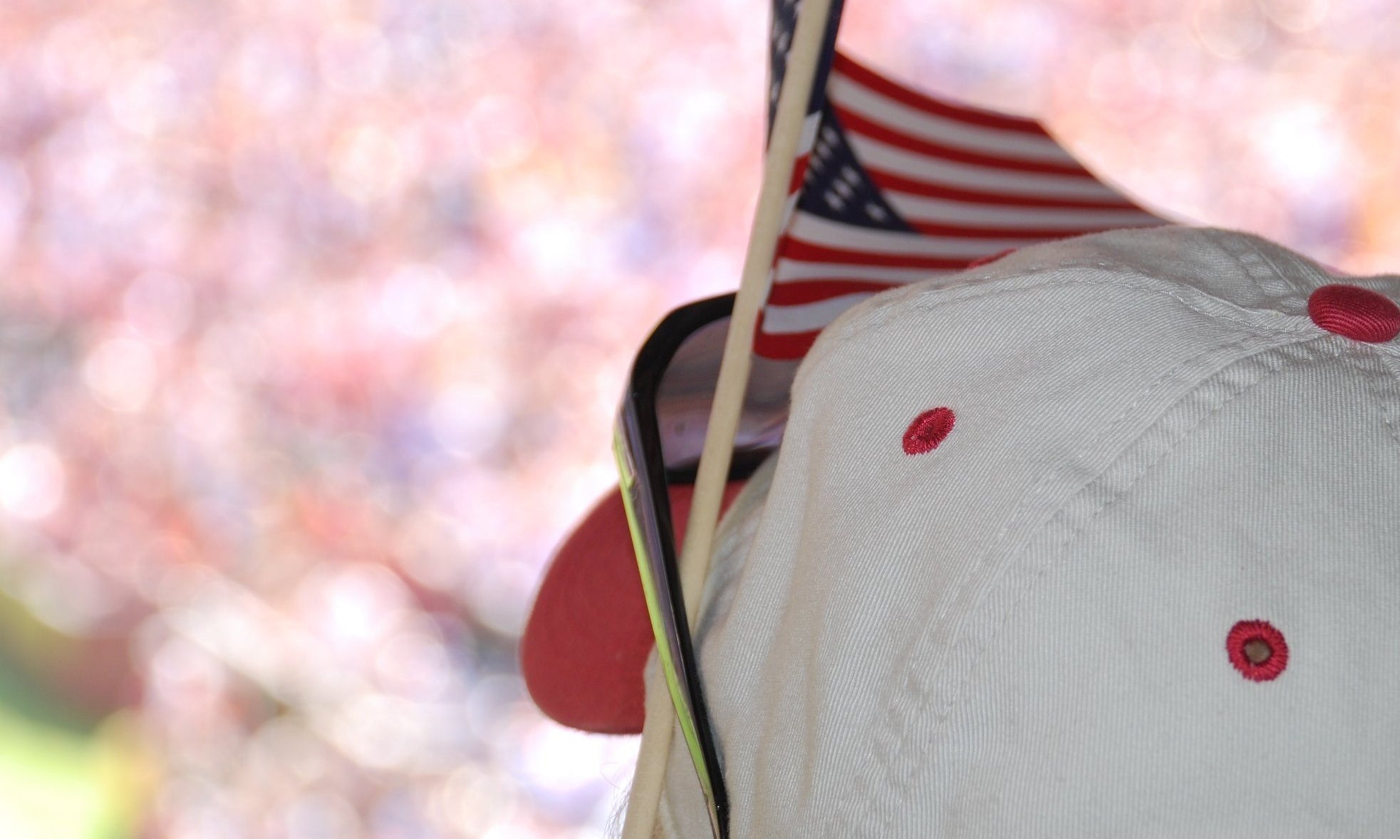 Person wearing a baseball hat with sunglasses resting on the brim and an American flag propped up between the hat and the sunglasses